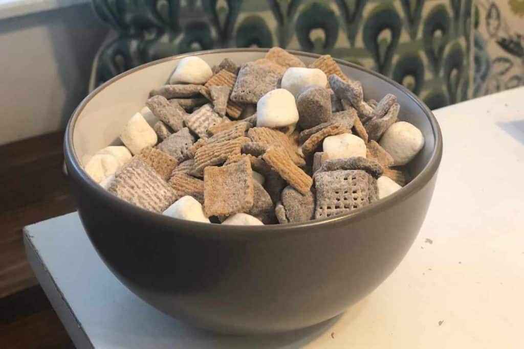 Gray bowl of Smores Snack Mix on a wooden table, including marshmallows, graham cereal and chocolate covered chex
