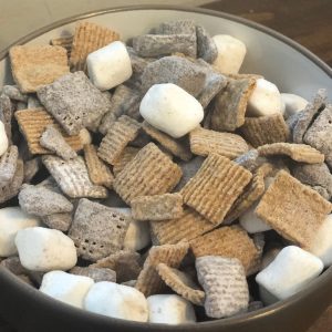 Gray bowl of Smores Snack Mix on a wooden table, including marshmallows, graham cereal and chocolate covered chex