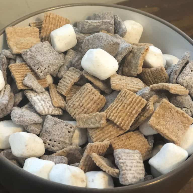 Gray bowl of Smores Snack Mix on a wooden table, including marshmallows, graham cereal and chocolate covered chex