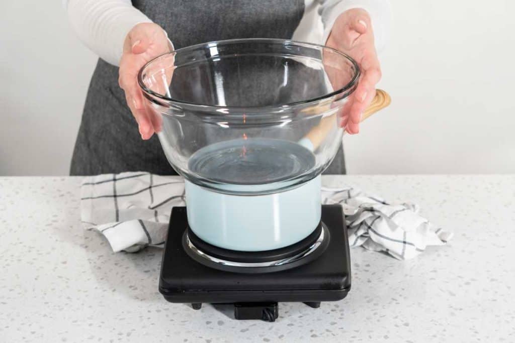 Induction cooking top with a saucepan and glass bowl as a double boiler on a kitchen countyer, with a lady in an apron holding the glass bowl in place