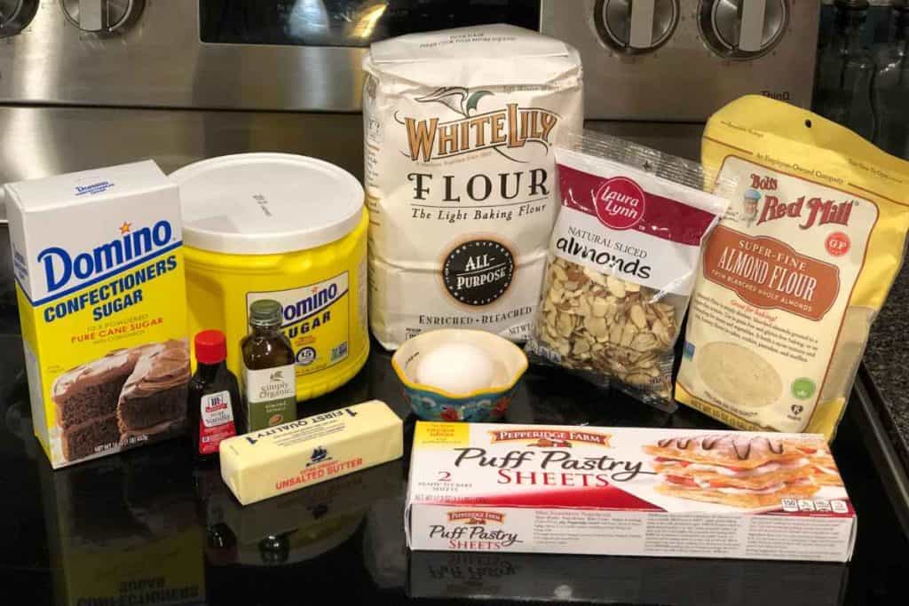 Ingredients for homemade almond croissants including almond flour, puff pastry, an egg, unsalted butter, sliced almonds, all-purpose flour, sugar, almond extract, vanilla extract and confectioner's sugar