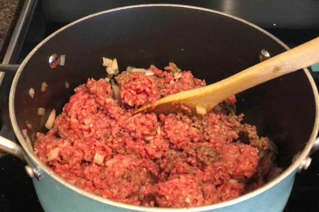 Ground beef cooking in a soup pot with chopped up onions in a blue pot