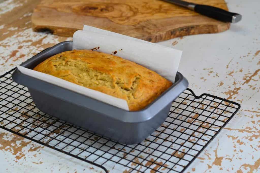 A loaf of banana bread fresh out of the oven in its loaf pan sitting on a wire cooling rack