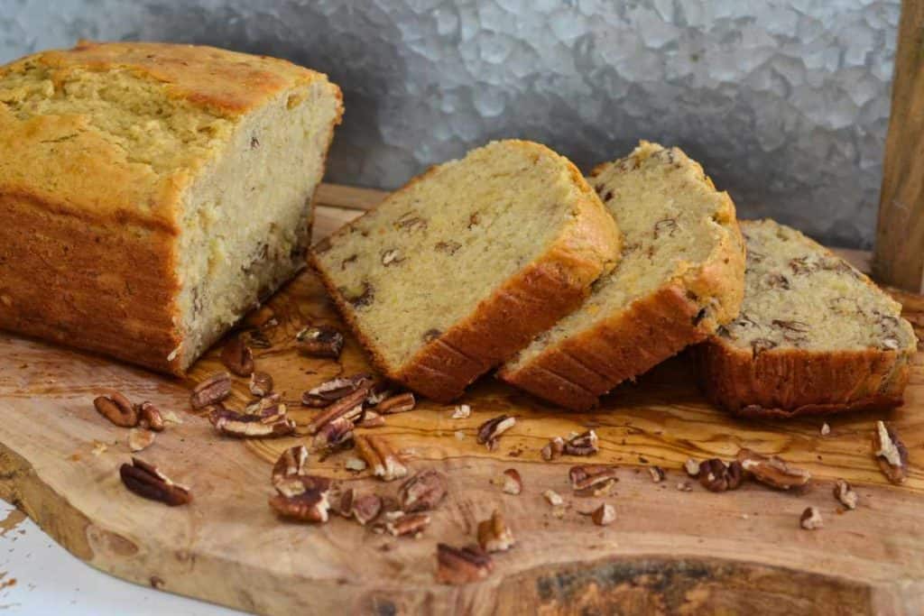 A loaf of banana bread with 3 slices cut and off to the side on a wooden cutting board with pecans sprinkled around