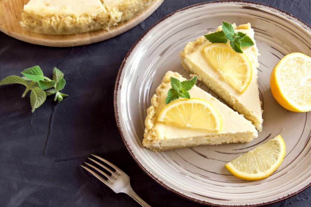 2 slices of no-bake lemonade cheesecake on white plates on a white tablecloth, with mint sprigs and lemon slices