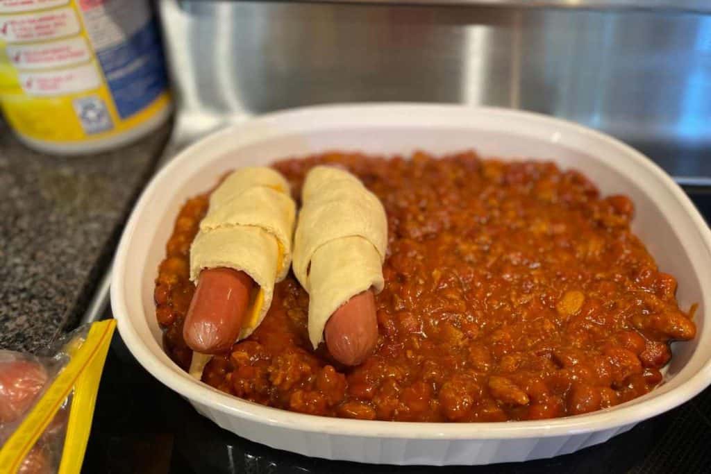 2 hot dogs wrapped with cheese and crescent rolls laying on top of a bed of chili