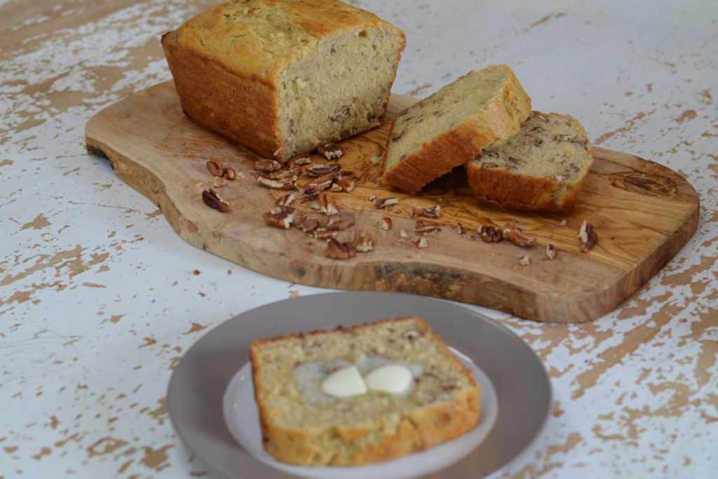 Loaf of banana bread halfway sliced sitting on a wooden cutting board on a white table with a plate alongside with a slice of banana bread and butter on it.