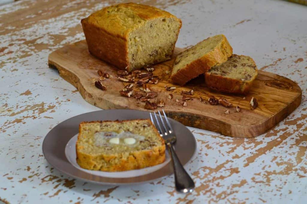 Loaf of banana bread halfway sliced sitting on a wooden cutting board on a white table with a plate alongside with a slice of banana bread and butter on it with a fork.