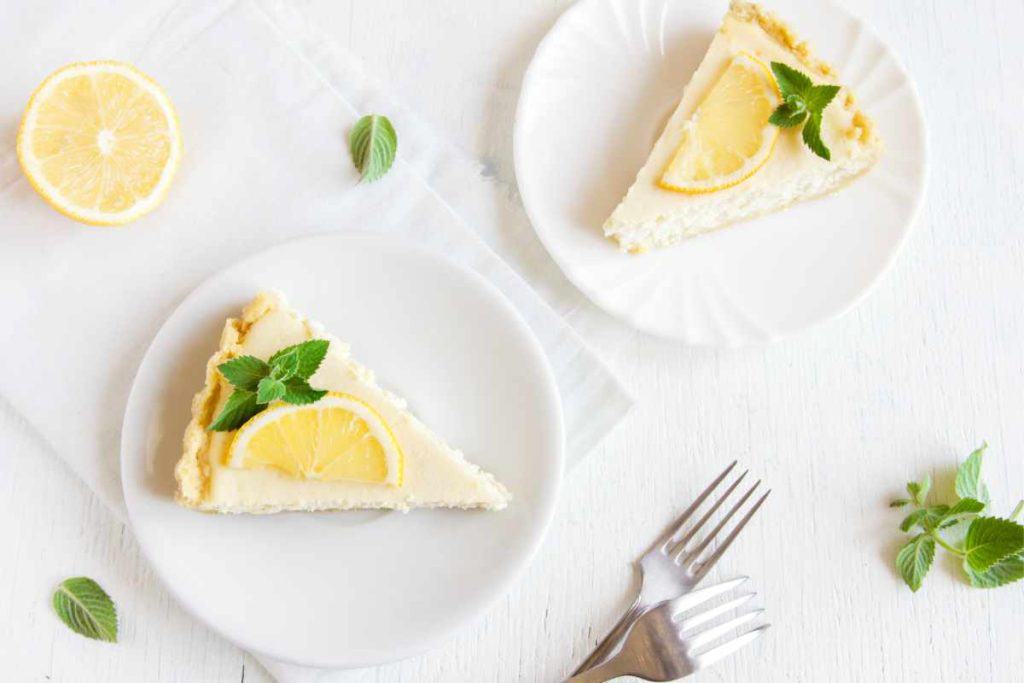 2 slices of no-bake lemonade cheesecake on white plates on a white tablecloth, with mint sprigs and lemon slices