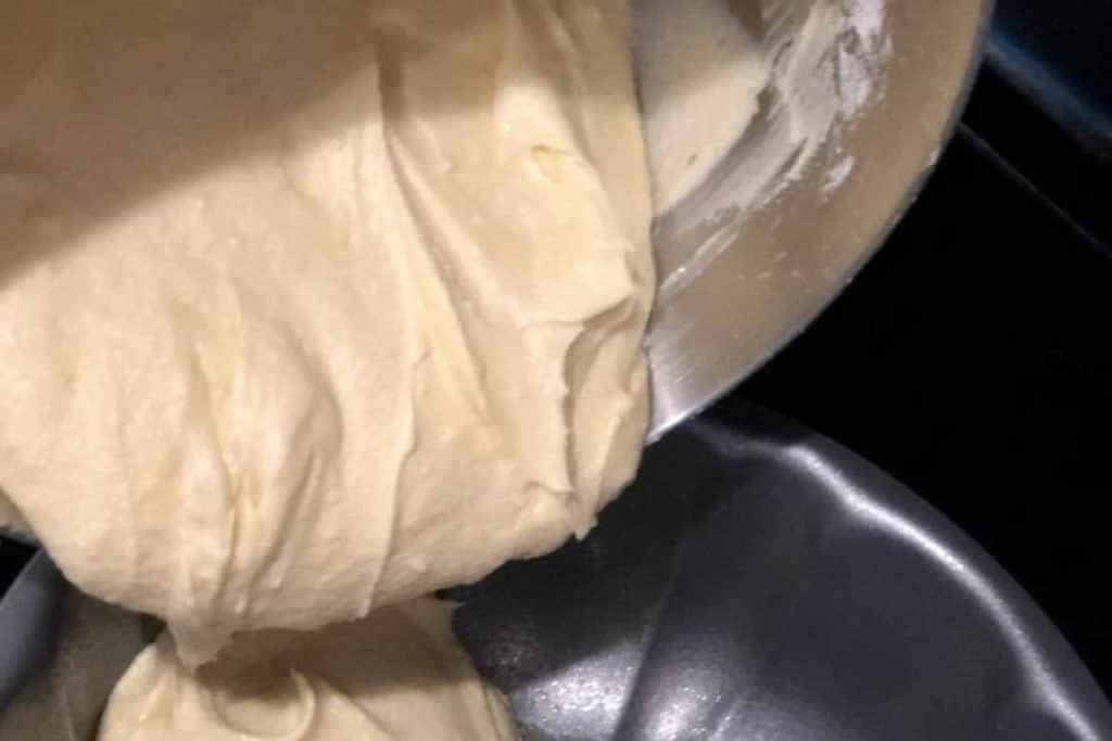 Lemon pound cake batter being poured from a stainless mixing bowl into a non-stick bundt cake pan