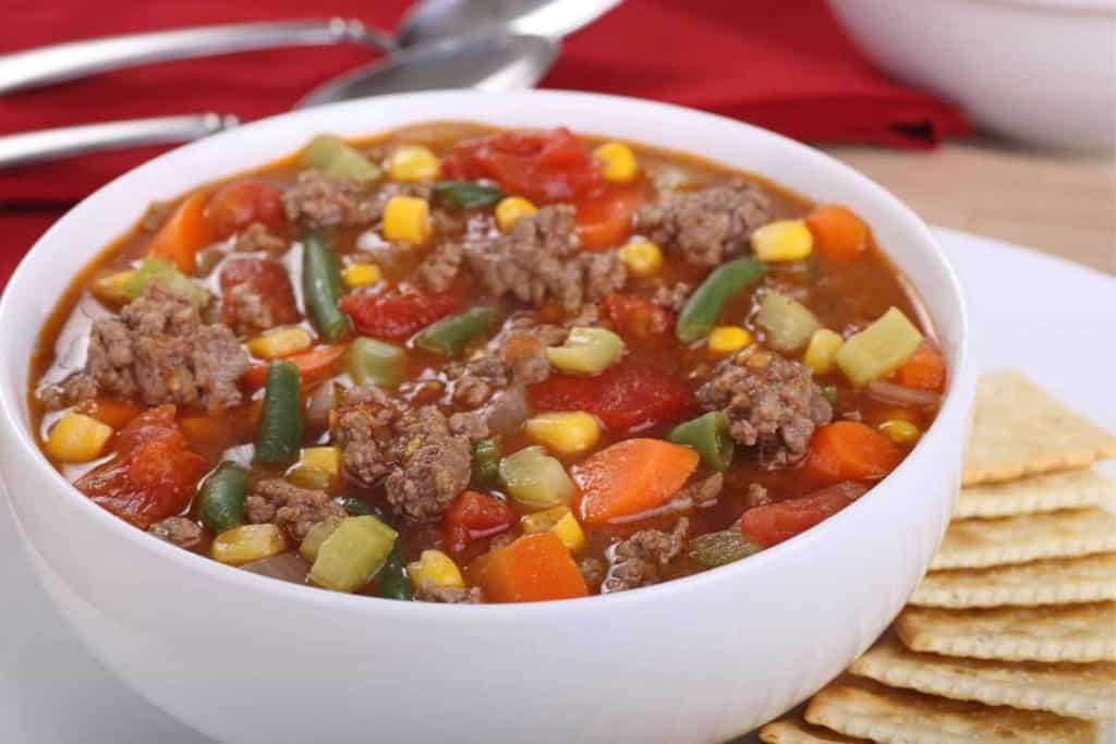 A white bowl of vegetable beef soup on a white plate with crackers on the plate, with a red napkin and spoon