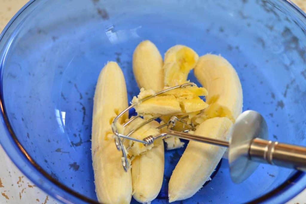 Pastry cutter mashing up bananas in a glass blue bowl for banana bread recipe