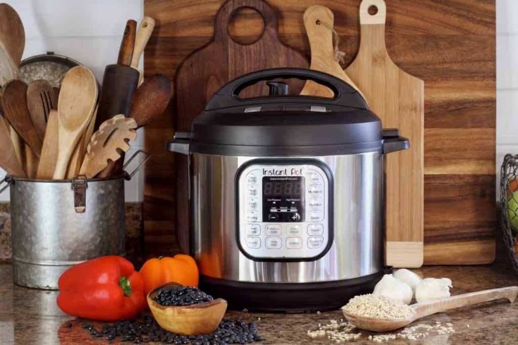 Instant pot sitting on the kitchen counter with wooden cutting boards and a crock of wooden utensils next to it along with dry rice and beans as well as some bell peppers on the counter.