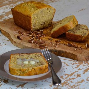 Loaf of banana bread halfway sliced sitting on a wooden cutting board on a white table with a plate alongside with a slice of banana bread and butter on it with a fork.