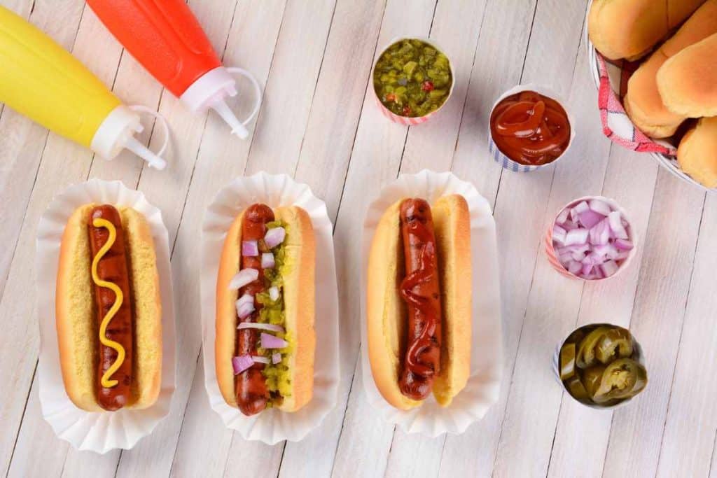 3 hot dogs with different condiments, and then all the hot dog condiments around the hot dogs in containers, including chopped onions, sliced jalepenos, ketchup, mustard and pickle relish