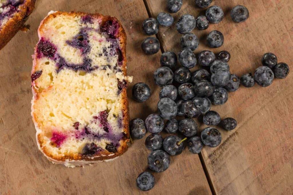 A slice of lemon blueberry loaf and some fresh blueberries on a wooden table