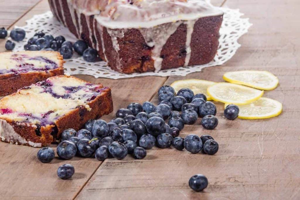 Lemon Blueberry Loaf on a paper doile on a wooden table, surrounded by slices of Lemon blueberry loaf and fresh blueberries and sliced lemons