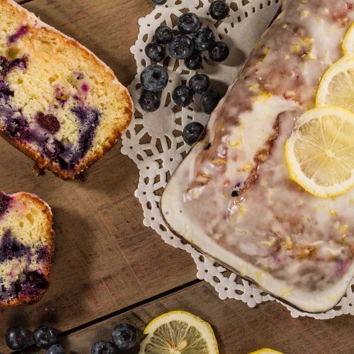 Blueberry Lemon Loaf on a paper doile with slices of the loaf and blueberries and sliced lemons all around the wooden table