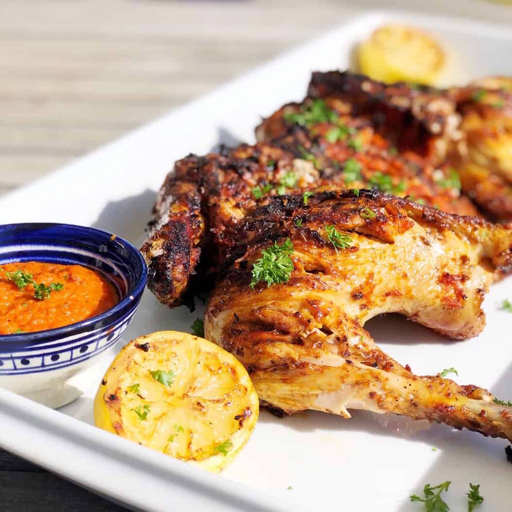This is a large white platter filled with Portuguese Chicken Piri Piri, which is grilled chicken and a red sauce in a bowl on the side, with grilled lemons on the platter as well