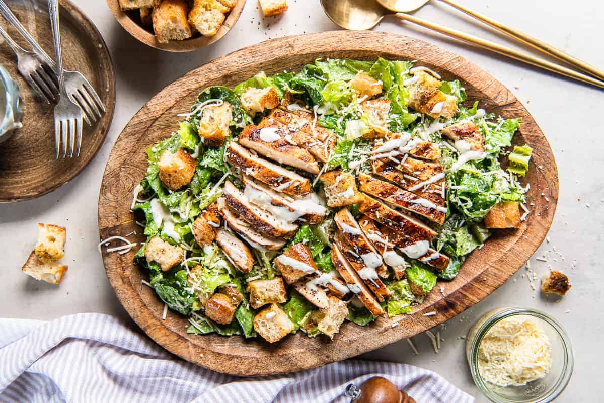 Grilled Chicken Caesar Salad on a wooden platter with croutons and forks nearby.