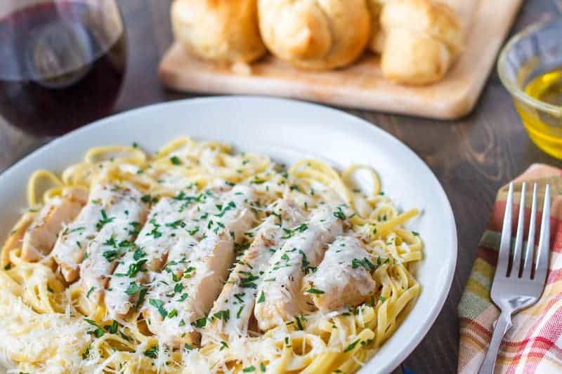 White bowl filled with fettucine alfredo pasta with grilled chicken on top covered with cheese and parsley