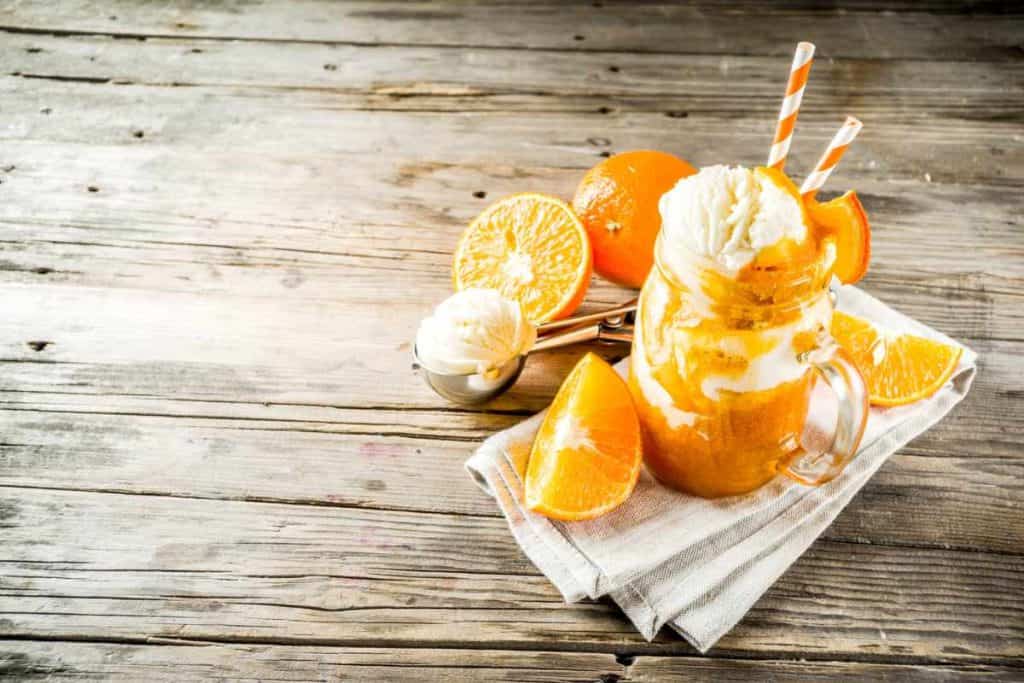 Orange cream ice cream float in a mason jar with a handle and cut up pieces of oranges arranged around the glass, along with a scoop of vanilla ice cream