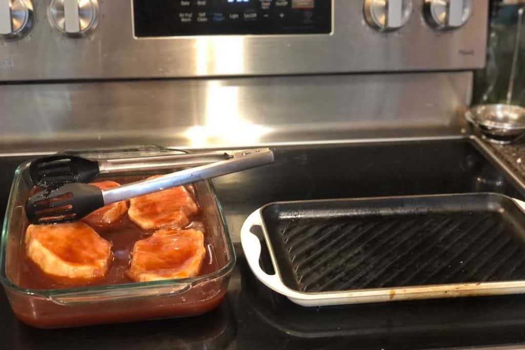 Marinading pork chops in a baking dish with bbq sauce marinade and tongs with a grill pan heating on the stove