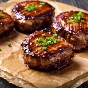 Grilled boneless pork chops covered in bbq sauce, on brown crinkled parchment paper with fresh chives as a garnish
