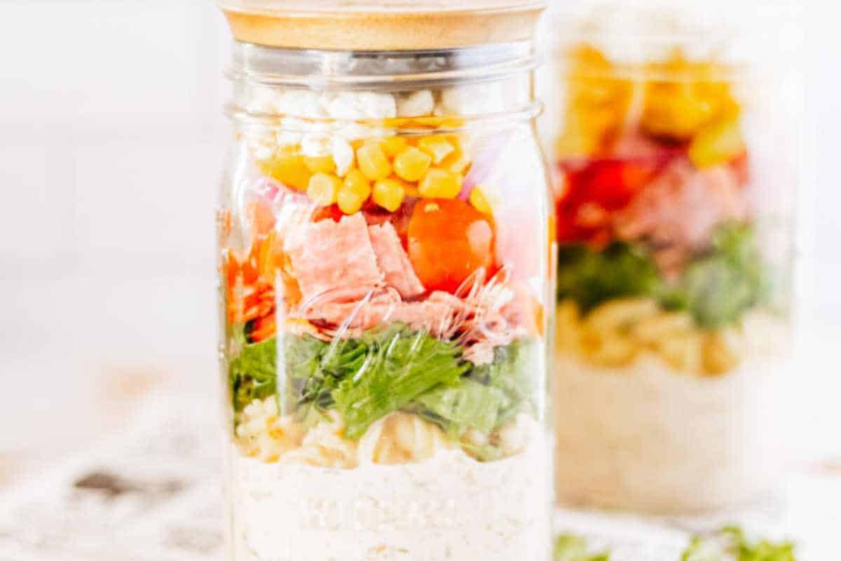 Italian Pasta Salad in a mason jar with the ingredients layered with the dressing on the bottom, then pasta, lettuce, salami, tomatoes, corn and cheese.