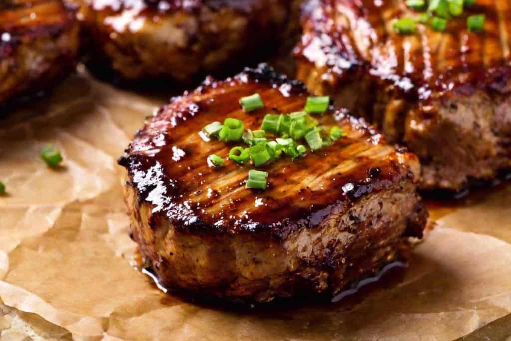 Grilled boneless pork chops covered in bbq sauce, on brown crinkled parchment paper with fresh chives as a garnish