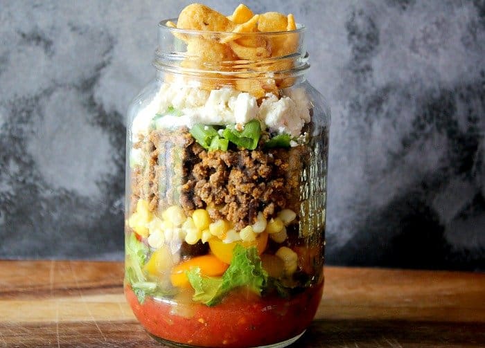 Taco salad layered in a mason jar, starting with salsa on the bottom, then tomatoes and lettuce, corn, seasoned beef, green onions, cheese and corn chips.