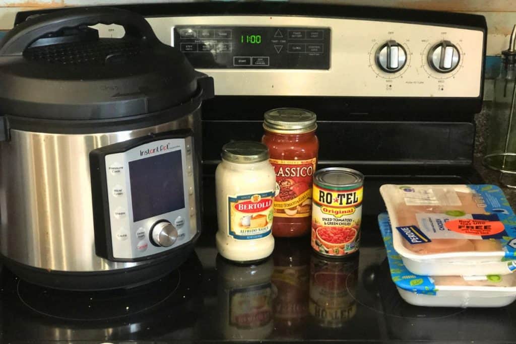 Ingredients and equipment for 3-Jar Slow-Cooker Creamy Tomato Chicken including a slow cooker, a jar of alfredo sauce, a jar of marinara sauce, a can of Rotel chopped tomatoes and two small packages of chicken breast