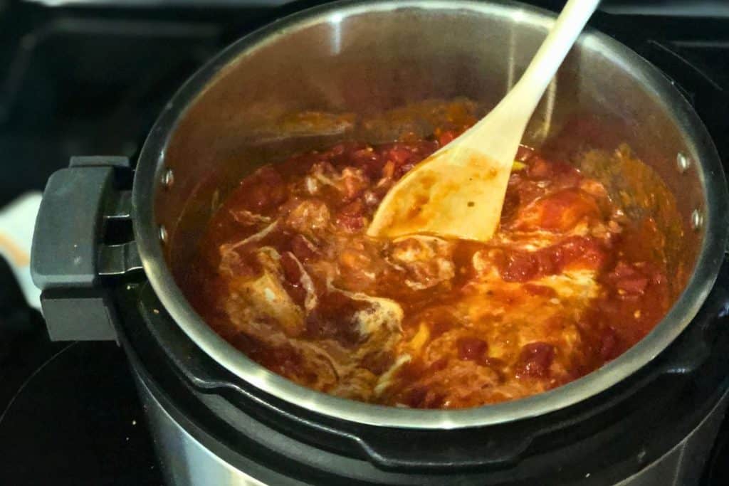 All ingredients for 3-Jar Slow-Cooker Creamy Tomato Chicken in a crock pot, but nothing is mixed together yet and there is also a wooden spoon in the pot