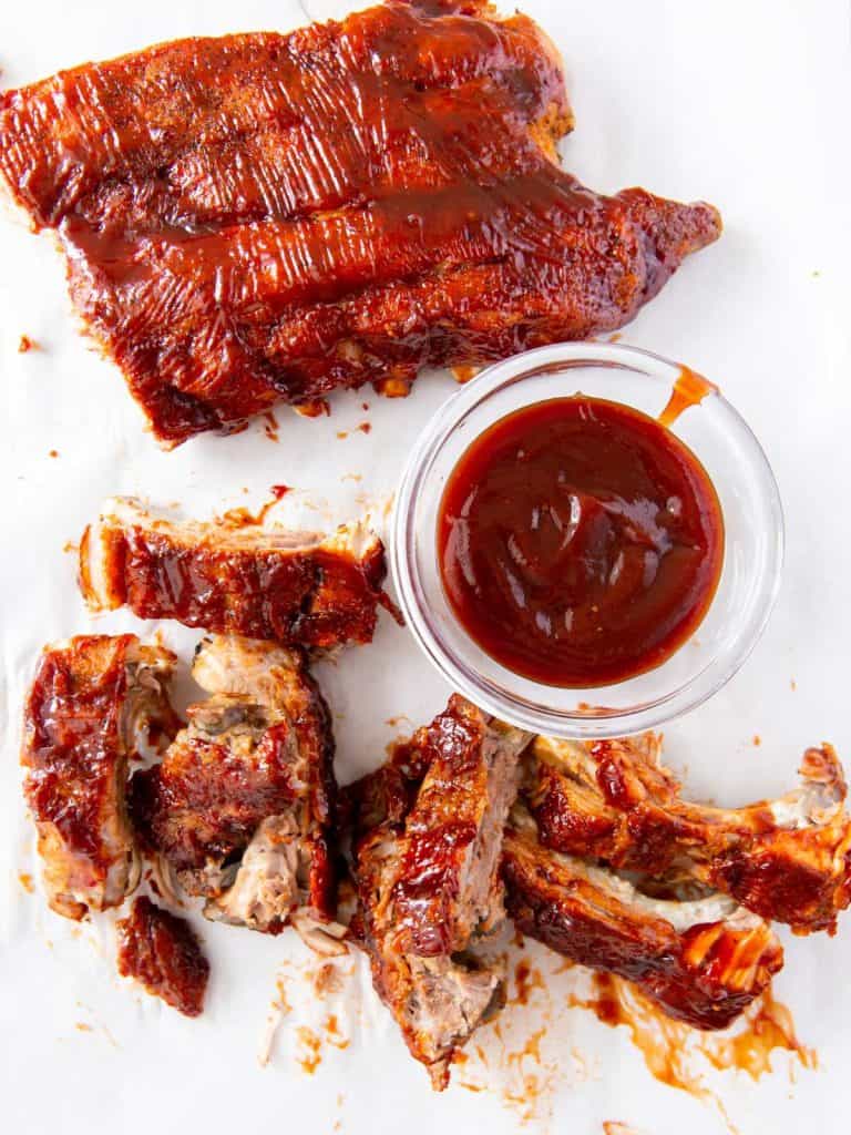 A rack of baby back ribs along with some cut up baby back ribs and a bowl of bbq sauce on a white tablecloth