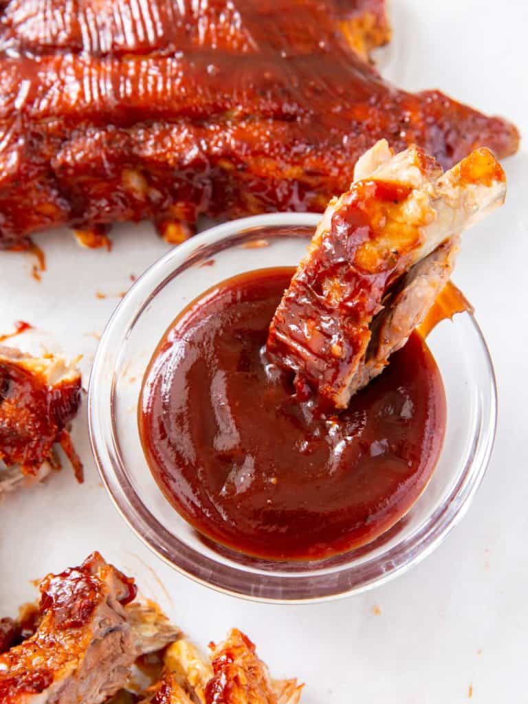 A baby back rib falling off the bone with tender meat dipping into a bowl of bbq sauce with other ribs all around the bowl