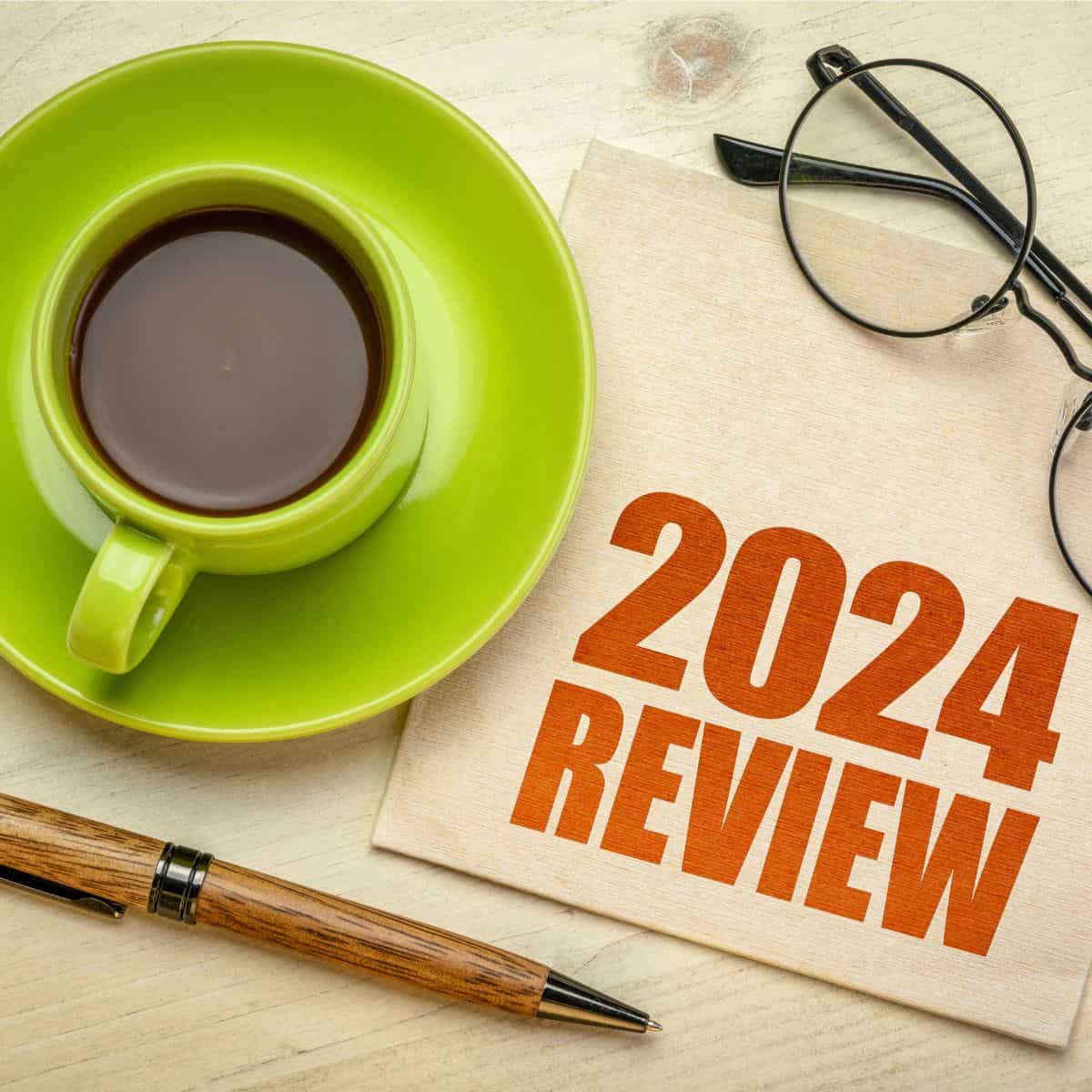 Green cup of coffee on a green saucer with a napkin that says "2024 Review" and a pair of glasses and a pen