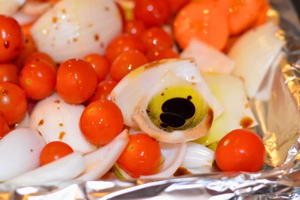 Balsamic vinegar drizzled over onions and tomatoes on a baking sheet getting ready to roast in the oven