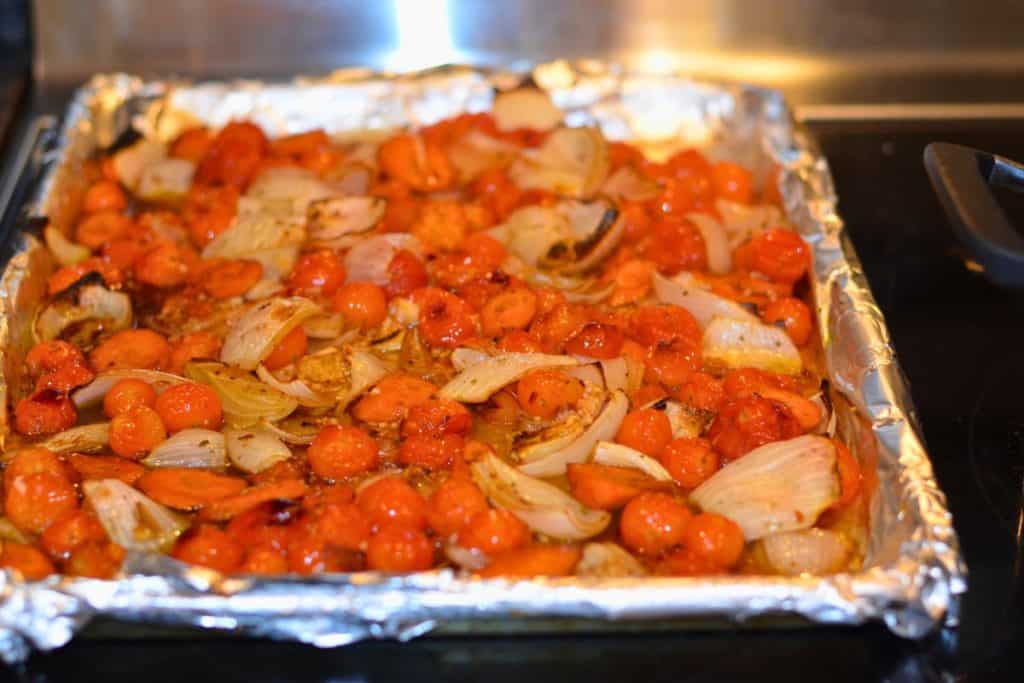 Roasted vegetables pulled from the oven on a foil lined baking sheet, including cherry tomatoes, carrots and onions