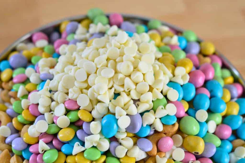 White chocolate chips on top of chocolate candies being added to the Easter Bunny Snack Mix recipe
