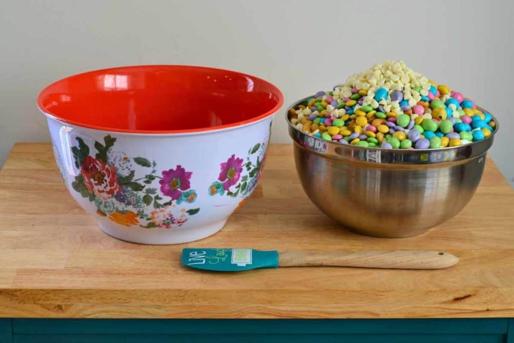 A stainless bowl full of unmixed Easter snack mix next to a larger empty bowl. There's also a spatula lying on the counter in front of hte bowls.