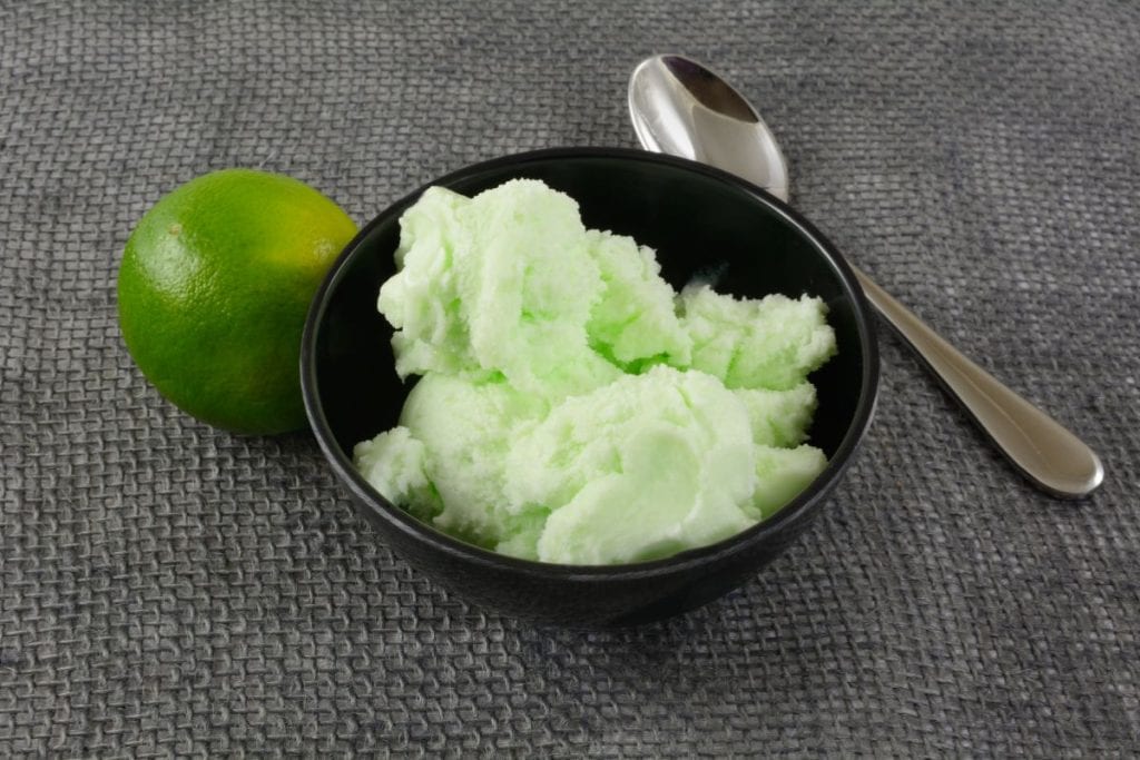 A bowl of the lime kool-aid ice cream with a spoon and a lime in the background of the picture