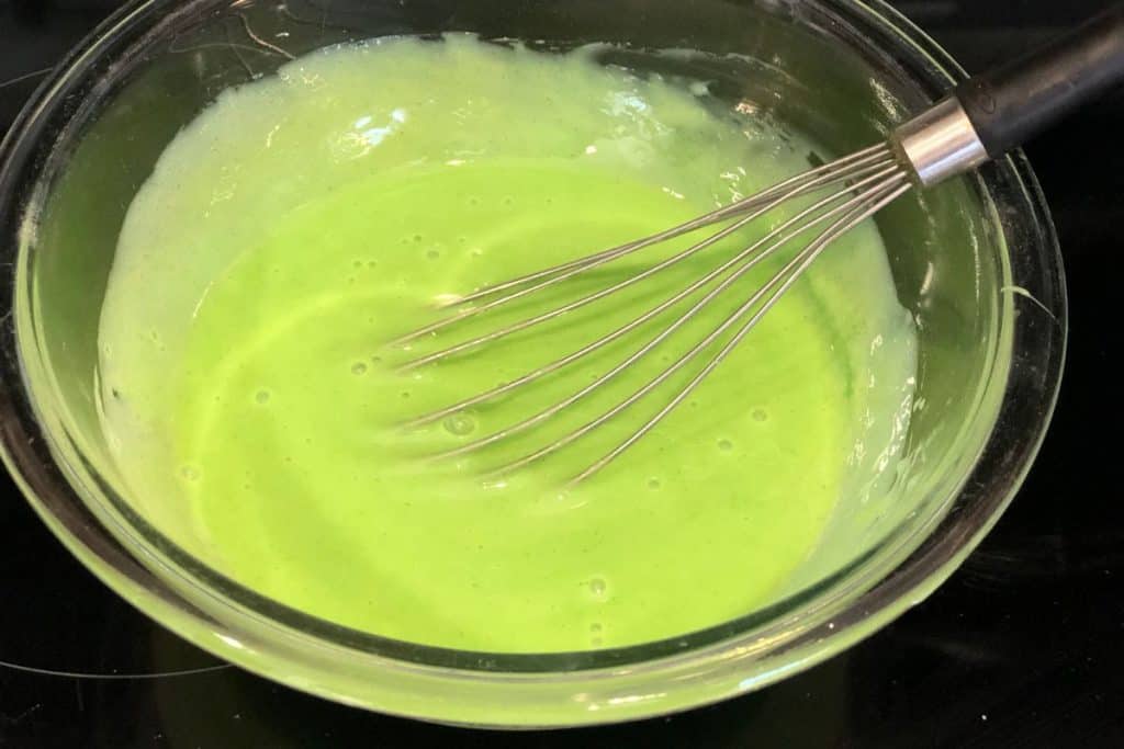 A glass bowl with sweetened condensed milk and lime kool aid mixed together with a flat whisk.