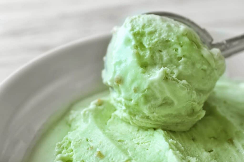 An ice cream scooper getting a scoop of lime kool aid ice cream from the bowl.