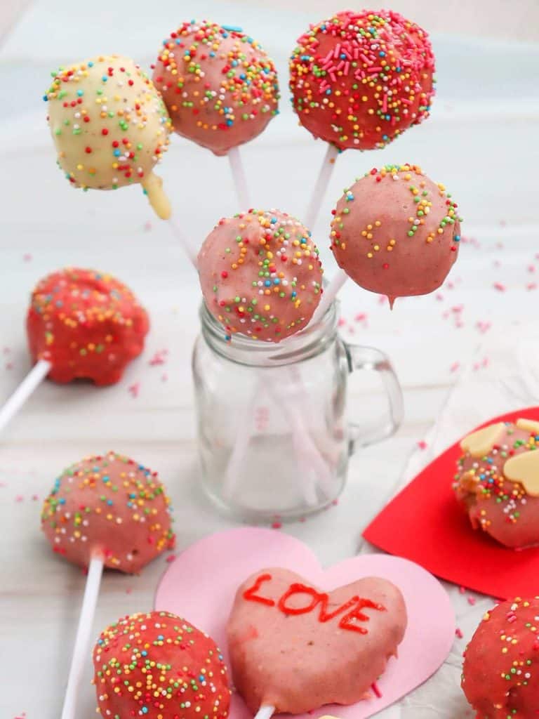A bouquet of Valentine's Cake pops in multiple shades of pink sitting upright in a jar with several of the cake pops also lying flat on the table around the jar.