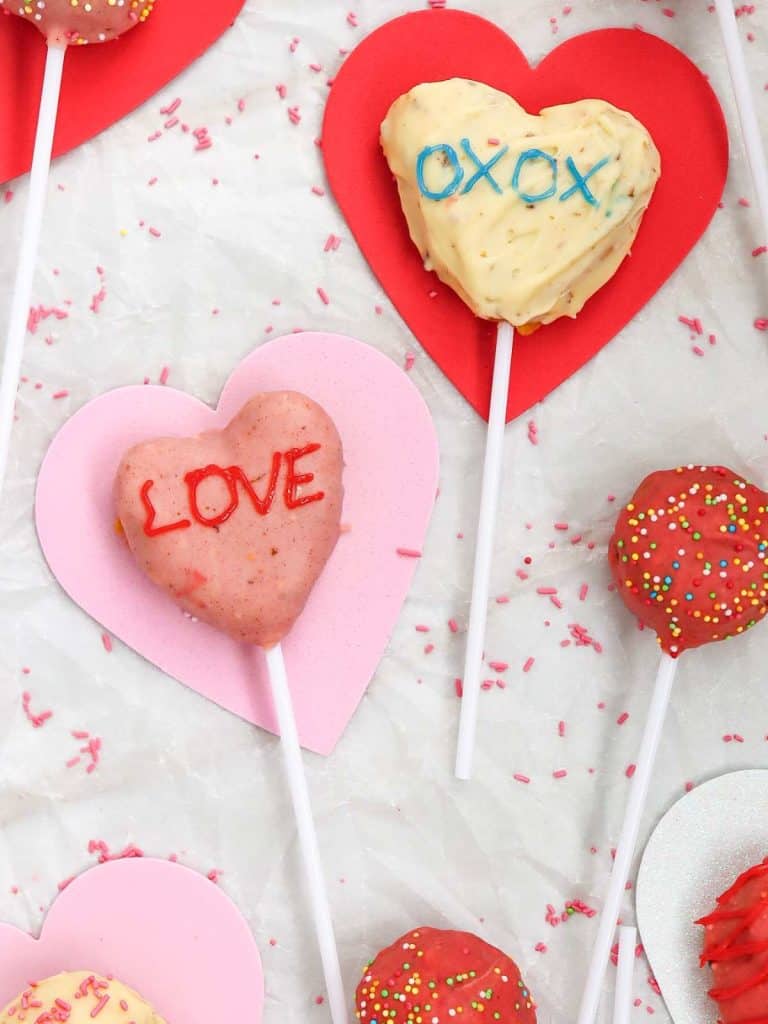 Two of the cake pops that are finished in a heart shape, one white and one pink lying flat on a piece of parchment paper with cut out hearts
