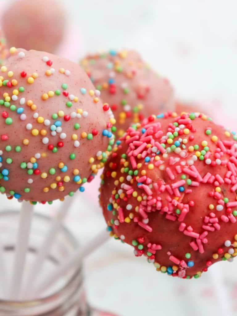 A close up of three of the cake pops in various shades of pink that have all colors of sprinkles on them for decoration. The pops are sitting upright in a jar.