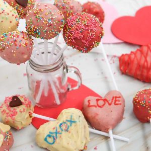 A bouquet of cake pops in a mason jar with other decorated cake pops lying flat on the table all around the jar, along with paper hearts and sprinkles.