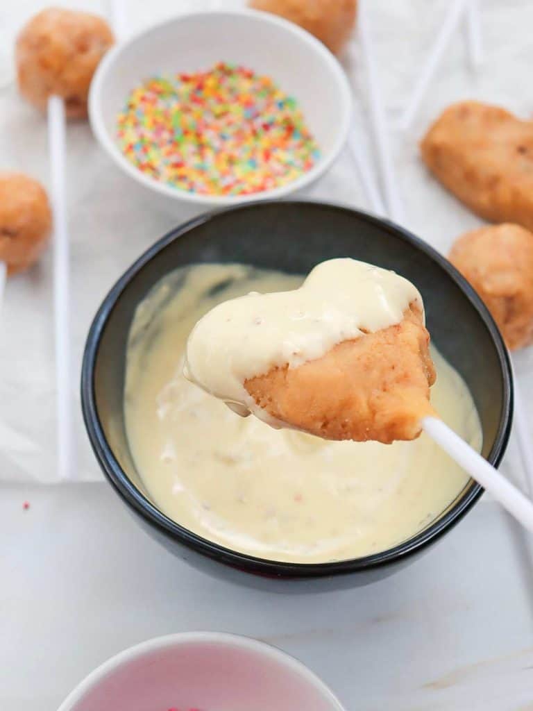 A heart-shaped cake pop being held over the bowl of white chocolate, halfway dunked and coated in white chocolate.