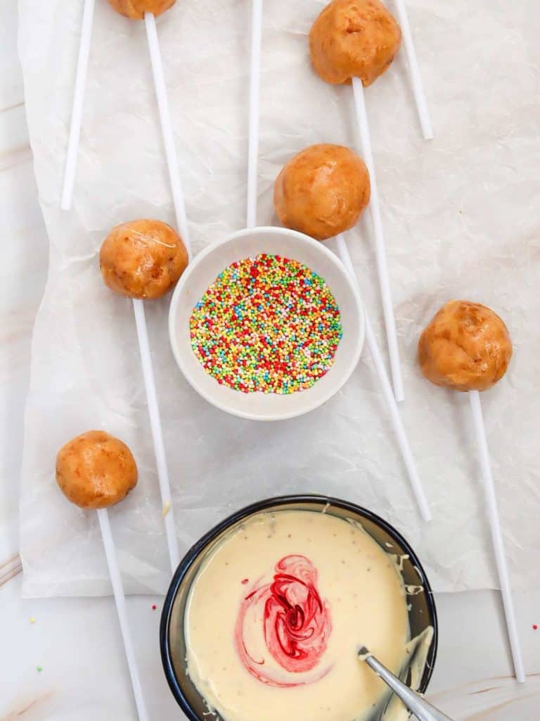 A bowl of sprinkles and a bowl of white chocolate that has food coloring just put into it. Also there are several raw cake pops ready to be coated lying on parchment paper around the two bowls.