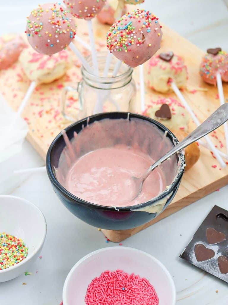 Melted white chocolate in a bowl that has pink food coloring added in to make the coating pink for the Valentine's Day Cake Pops. In the background are some already finished cake pops standing in a mason jar along with all the sprinkles and decorations for the pops